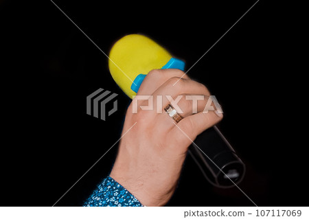A man's hand close-up holds a microphone with a yellow soft rim, a karaoke singing tool on a dark background A man's hand close-up holds a microphone with a yellow soft rim, a karaoke singing tool on a dark background 107117069