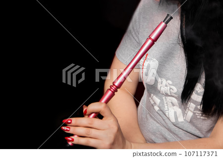 The hand of a girl with a red manicure holds a hookah pipe, a smoking object on a dark background The hand of a girl with a red manicure holds a hookah pipe, a smoking object on a dark background 107117371