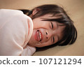 Cute Asian girl lying on the wooden floor at home smiling happily at the camera 107121294