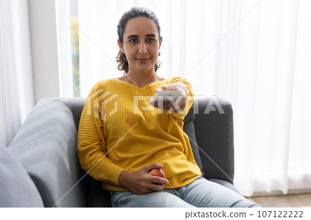Woman sitting comfortable at home sofa watching tv pointing remote control for changing tv channel program 107122222