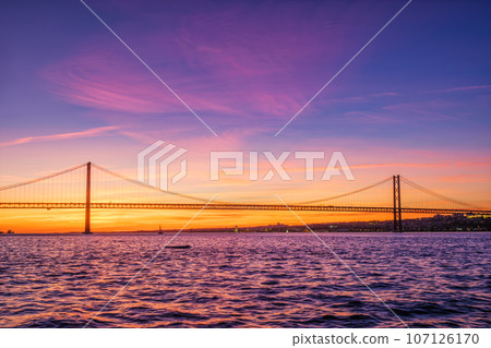 View of 25 de Abril Bridge famous tourist landmark of Lisbon connecting Lisboa and Almada over Tagus river with tourist yacht silhouette at sunset. Lisbon, Portugal View of 25 de Abril Bridge famous tourist landmark of Lisbon connecting Lisboa and Almada over Tagus river with tourist yacht silhouette at sunset. Lisbon, Portugal 107126170