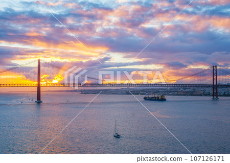 View of 25 de Abril Bridge famous tourist landmark of Lisbon over Tagus river with tourist yacht boats and cargo container ship on sunset. Lisbon, Portugal View of 25 de Abril Bridge famous tourist landmark of Lisbon over Tagus river with tourist yacht boats and cargo container ship on sunset. Lisbon, Portugal 107126171