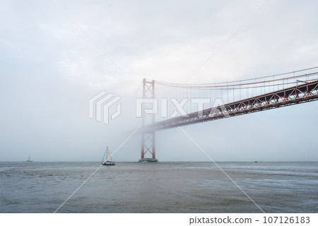 View of 25 de Abril Bridge famous tourist landmark of Lisbon connecting Lisboa and Almada in heavy fog mist wtih yacht boats passing under. Lisbon, Portugal View of 25 de Abril Bridge famous tourist landmark of Lisbon connecting Lisboa and Almada in heavy fog mist wtih yacht boats passing under. Lisbon, Portugal 107126183