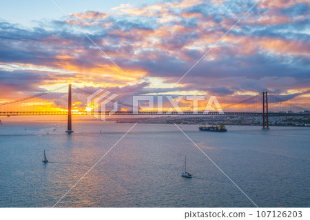 View of 25 de Abril Bridge famous tourist landmark of Lisbon over Tagus river with tourist yacht boats and cargo container ship on sunset. Lisbon, Portugal View of 25 de Abril Bridge famous tourist landmark of Lisbon over Tagus river with tourist yacht boats and cargo container ship on sunset. Lisbon, Portugal 107126203