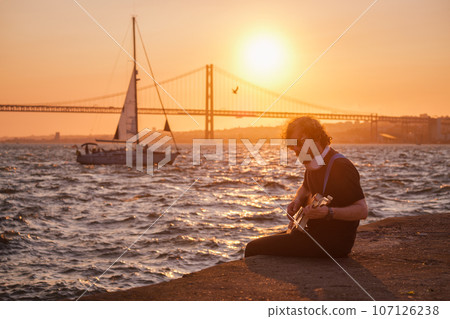 Hipster street musician in black playing electric guitar in the street on sunset on embankment with 25th of April bridge and yacht boat in background. Lisbon, Portugal Hipster street musician in black playing electric guitar in the street on sunset on embankment with 25th of April bridge and yacht boat in background. Lisbon, Portugal 107126238