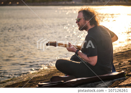 Hipster street musician in black playing electric guitar in the street sitting on pier embankment on sunset Hipster street musician in black playing electric guitar in the street sitting on pier embankment on sunset 107126261