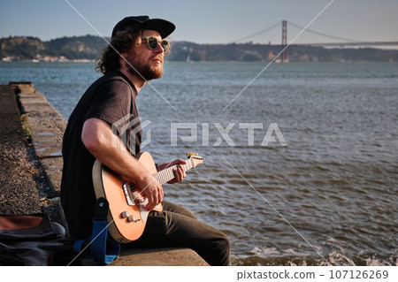 Hipster street musician in black playing electric guitar in the street sitting on pier embankment on sunset in Lisbon, Portugal 107126269