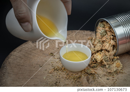Pouring Chinese chrysanthemum flower tea from the small jug into a cup. Dried chrysanthemum flower is on wooden cutting board. Healthy beverage for drink. Pouring Chinese chrysanthemum flower tea from the small jug into a cup. Dried chrysanthemum flower is on wooden cutting board. Healthy beverage for drink. 107130673