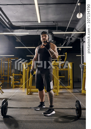 Attractive, tattooed, bearded man in black sport gloves, shorts, vest, cap and sneakers. Showing cool sign, barbell is on floor. Dark gym. Full length 107134790