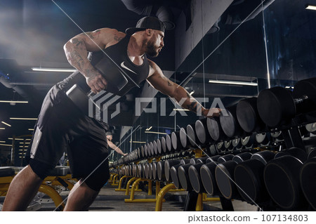 Tattooed, muscular guy in black shorts, vest and cap. Exercising with dumbbells for training his triceps in dark gym. Sport, fitness. Close up 107134803
