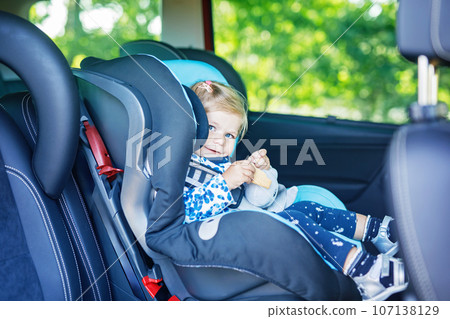 Adorable baby girl with blue eyes sitting in car safety seat. Toddler child going on family vacations and jorney. Smiling happy child during traffic jam, eating bisquit 107138129