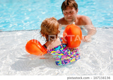 Cute happy little toddler girl and father in the pool and having fun on family vacations in a hotel resort. Healthy child and man playing in water. Baby daughter in colorful fashion swimsuit. 107138136