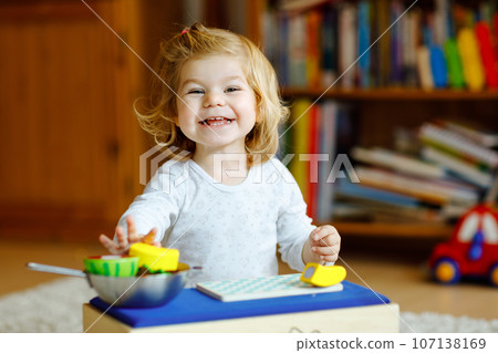 Cute little toddler girl playing at home with eco wooden toys. Happy healthy excited child cutting vegetables and fruits with toy knife. Baby girl having fun with role kitchen and cooking game. 107138169