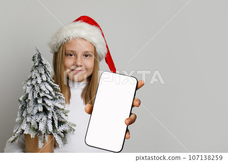 Christmas portrait of cute kid wearing Santa hat holds smartphone with blank empty screen display 107138259