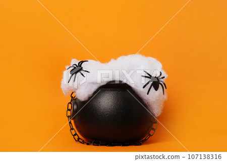 Happy Halloween holiday concept. Witch's cauldron with black spiders on orange background, copy space. Witch's cauldron as a pedestal for products 107138316