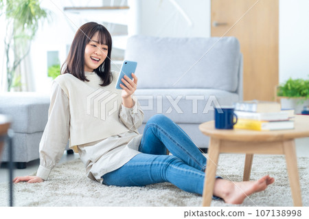 一位年輕女子在客廳一邊放鬆一邊看著智慧型手機 一位年輕女子在客廳一邊放鬆一邊看著智慧型手機 107138998
