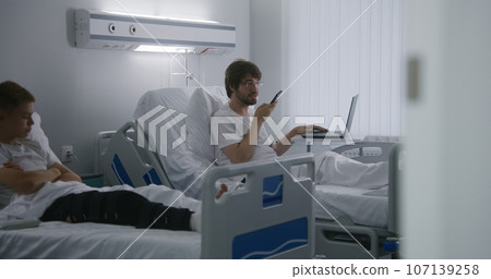 Man lies on bed and chats using phone in hospital Man lies on bed and chats using phone in hospital 107139258