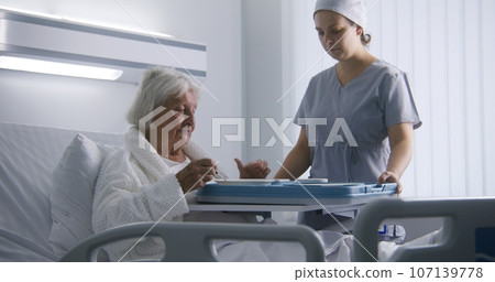 Nurse opens big lunch box with meal to female old patient Nurse opens big lunch box with meal to female old patient 107139778