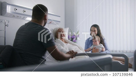 Female old patient lies in bed in hospital ward, spends time with relatives 107139846