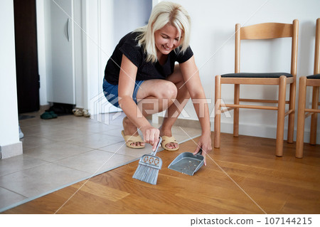 A young woman sweeps the floor of her home with a dustpan and broom. A young woman sweeps the floor of her home with a dustpan and broom. 107144215