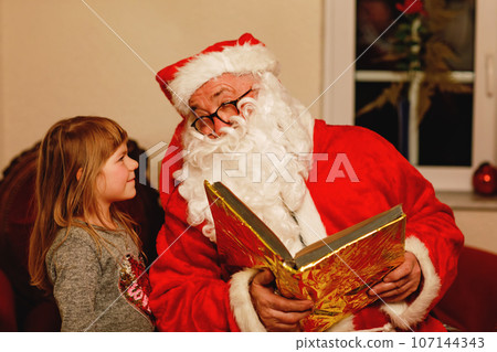 Little toddler girl talking to Santa Claus called Nikolaus or Weihnachtsmann in German. Happy smiling kid waiting for gifts. Santa with golden book. Xmas, childhood, holiday Little toddler girl talking to Santa Claus called Nikolaus or Weihnachtsmann in German. Happy smiling kid waiting for gifts. Santa with golden book. Xmas, childhood, holiday 107144343