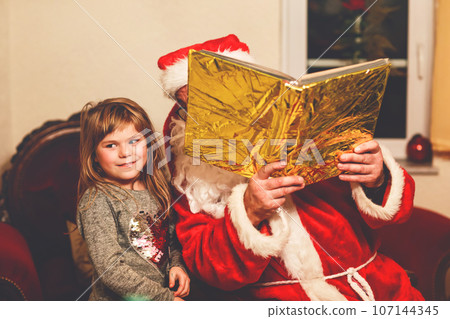 Little toddler girl talking to Santa Claus called Nikolaus or Weihnachtsmann in German. Happy smiling kid waiting for gifts. Santa with golden book. Xmas, childhood, holiday Little toddler girl talking to Santa Claus called Nikolaus or Weihnachtsmann in German. Happy smiling kid waiting for gifts. Santa with golden book. Xmas, childhood, holiday 107144345
