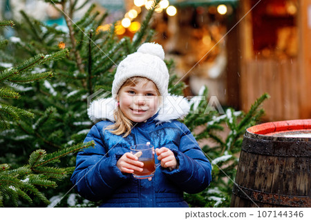 Little cute preschool girl drinking hot children punch or chocolate on German Christmas market. Happy child on traditional family market in Germany, Laughing boy in colorful winter clothes 107144346