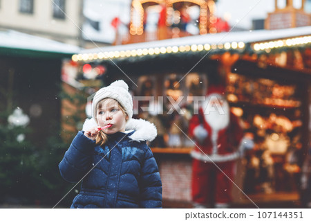 Little cute preschool girl with candy cane from a sweets stand on Christmas market. Happy child on traditional family market in Germany. Preschooler in colorful winter clothes during snowfall Little cute preschool girl with candy cane from a sweets stand on Christmas market. Happy child on traditional family market in Germany. Preschooler in colorful winter clothes during snowfall 107144351