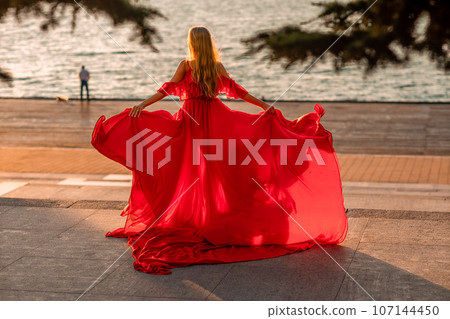 Sunrise red dress. A woman in a long red dress against the backdrop of sunrise, bright golden light of the sun's rays. The concept of femininity, harmony. 107144450