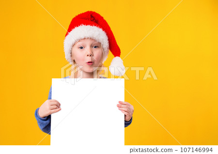 Cute little girl in Santa Claus hat with a white blank paper sheet. Christmas, x-mas, New Year, Winter, people, advertisement, sale concept. Cute little girl in Santa Claus hat with a white blank paper sheet. Christmas, x-mas, New Year, Winter, people, advertisement, sale concept. 107146994