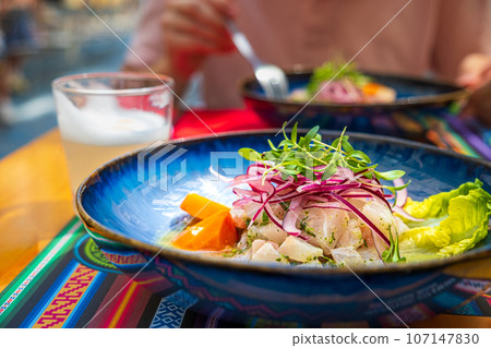 Fresh ceviche served in blue plate over placemat with traditional patterns, Pisco Sour cocktail. Unrecognizable adult in blurred background. Peruvian-style ceviche made out of raw fish, red onions Fresh ceviche served in blue plate over placemat with traditional patterns, Pisco Sour cocktail. Unrecognizable adult in blurred background. Peruvian-style ceviche made out of raw fish, red onions 107147830