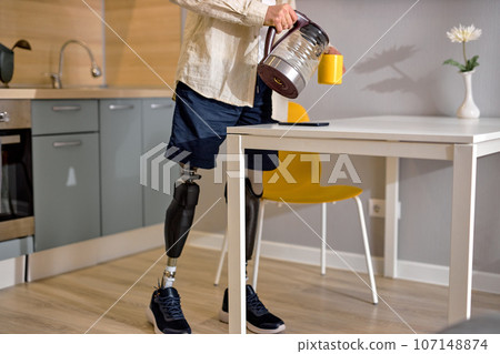 disabled guy with leg prosthesis in kitchen at home, making tea in the morning disabled guy with leg prosthesis in kitchen at home, making tea in the morning 107148874