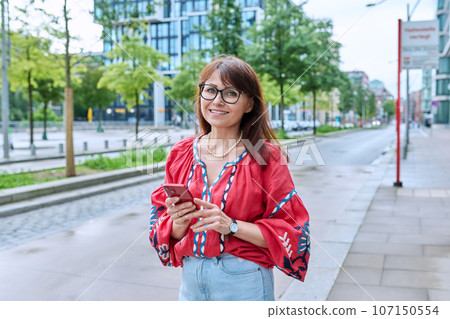 Mature smiling woman in embroidered shirt looking at camera, on street of modern city Mature smiling woman in embroidered shirt looking at camera, on street of modern city 107150554