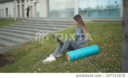 Happy woman, sitting laptop outdoor. Smiling fit girl typing on laptop computer at city background. Person working on laptop outside. Attractive girl, corporate startup office employee working outdoor 107152878
