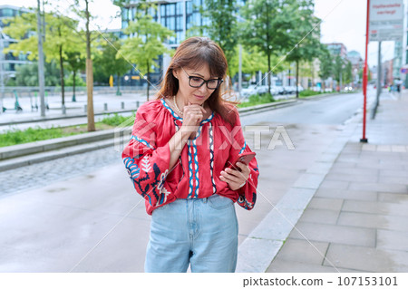 Mature smiling woman in embroidered shirt looking at smartphone, on street of modern city Mature smiling woman in embroidered shirt looking at smartphone, on street of modern city 107153101