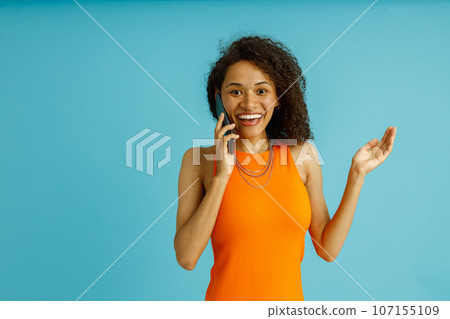 Positive woman in orange dress is talking by mobile phone while standing on blue studio background  107155109