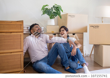 Happy young indian couple taking a break on moving day into new home sitting on floor surrounded by box 107155741