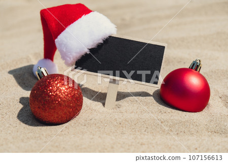 Empty blackboard frame board with copy space for your text or design displays on sandy beach. Christmas balls Santa hat New Year decoration. Summer vacation and holiday business travel concept 107156613