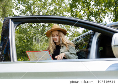 Smiling young woman in hat stoped on road to check the route on travel map. Local solo travel on weekends concept. Exited woman explore freedom outdoors in forest. Unity with nature lifestyle, rest 107156634