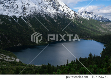 Morskie Oko Snowy Mountain Hut in Polish Tatry mountains, drone view, Zakopane, Poland. Aerial view shot of beautiful green hills and mountains in dark clouds and reflection on the lake Morskie Oko 107156749