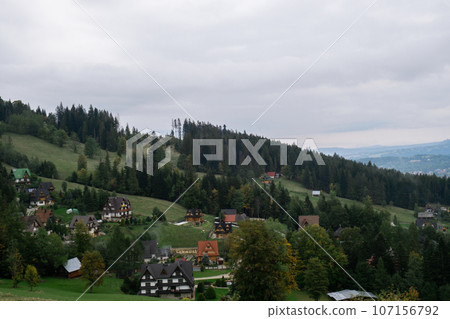 Aerial view from drone Picturesque landscape with small village with typical residential houses on grassy meadow surrounded by green and snowy mountains against cloudy blue sky in Zakopane Poland Aerial view from drone Picturesque landscape with small village with typical residential houses on grassy meadow surrounded by green and snowy mountains against cloudy blue sky in Zakopane Poland 107156792