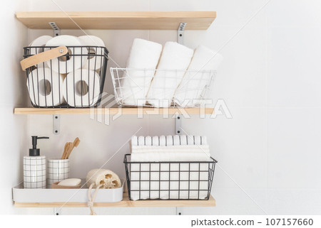 Beautiful white bathroom. Wooden shelves. Rolled towels, stacked towels and baskets. Soap, washcloth, toilet paper and toothbrushes. Indoor flowers. 107157660