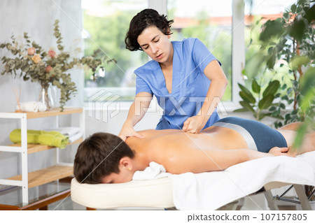 Adult woman giving back massage to young guy Adult woman giving back massage to young guy 107157805