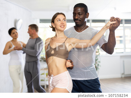 Positive African american man dancing playful Latin dance bachata with female partner during group class in choreography studio 107158234
