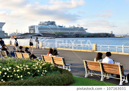 A couple sitting on a bench in Yamashita Park and a large cruise ship anchored at the main pier. 107163031