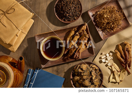 A wooden dish in rectangle-shaped featured a bowl of tonic and Red ginseng. Other herbal medicine types are surrounded with books, earthen pot and medicine packs A wooden dish in rectangle-shaped featured a bowl of tonic and Red ginseng. Other herbal medicine types are surrounded with books, earthen pot and medicine packs 107165871