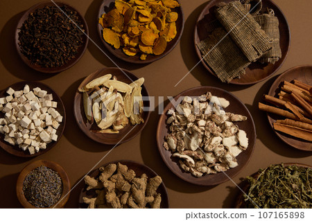 Round-shaped dishes of Bai zhu, Eucommia bark, Dried turmeric, Sand ginger, Lavender and other traditional medicine over the brown background. Medicine advertising Round-shaped dishes of Bai zhu, Eucommia bark, Dried turmeric, Sand ginger, Lavender and other traditional medicine over the brown background. Medicine advertising 107165898