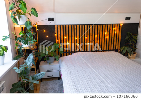 Loft style bedroom interior, black wall with wooden slats, metal bed, retro light bulbs garland, potted plants on a trapezoidal window in the attic. Modern Green House Loft style bedroom interior, black wall with wooden slats, metal bed, retro light bulbs garland, potted plants on a trapezoidal window in the attic. Modern Green House 107166366