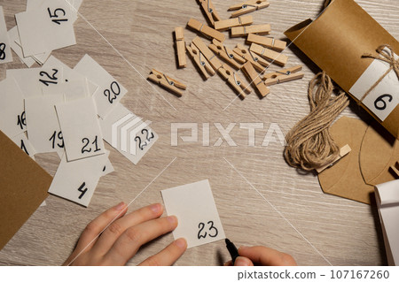 Unrecognizable young woman sticks number on craft bag, fastens with clothespin. Female making kraft paper for homemade advent calendar Made with your own hands step by step DIY crafts do it yourself Unrecognizable young woman sticks number on craft bag, fastens with clothespin. Female making kraft paper for homemade advent calendar Made with your own hands step by step DIY crafts do it yourself 107167260