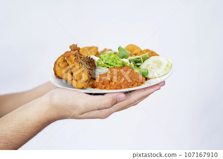 A human hand holding a plate with pecel ayam, fried tempe, and tofu 107167910